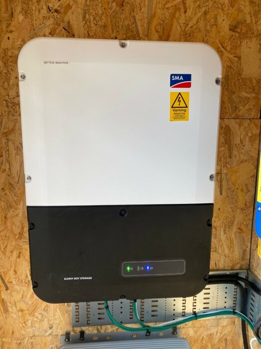 Bild på installationen av SMA Sunny Boy Storage 3.7 batteri-inverter monterad på en vägg i ett fritidshus, med varningsskylt och anslutande kablar.