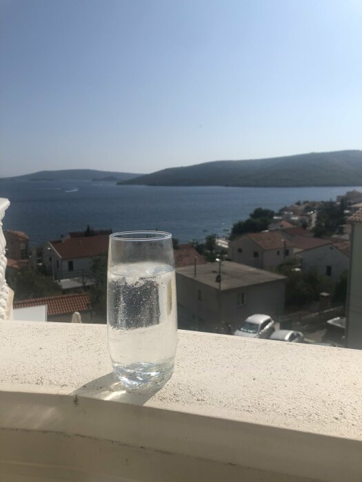 Ett glas gin och tonic står på en balkongräcke med utsikt över havet och hus i bakgrunden.