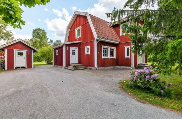 En röd fasad på ett nyinköpt hus som ska målas om med Falu Vapen Fasad; grön trädgård och garage syns i bakgrunden, en grusinfart leder till huset.