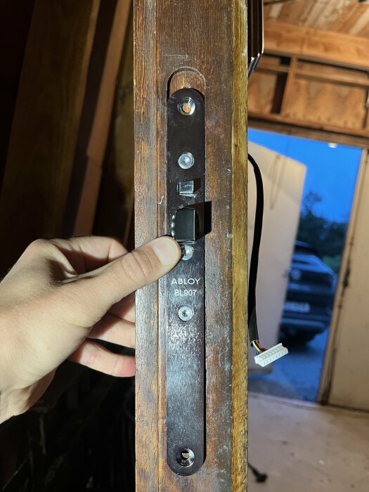 En hand håller i en dörrkarm med ett infällt lås märkt "ABLOY BL907" monterat i en träram, bredvid en kopplingskabel, inomhus i ett garage eller arbetsutrymme.