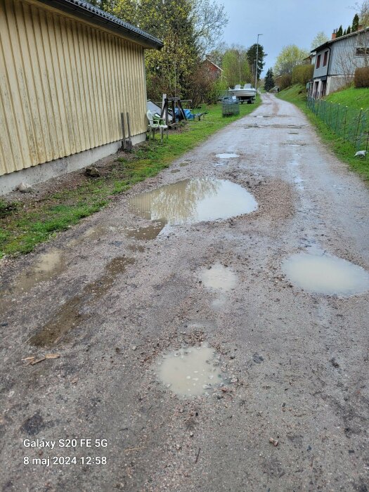 Grusväg med flera vattenpölar efter regn, med trädgårdsstolar och redskap längs en gul husvägg till vänster och bostadshus i bakgrunden.