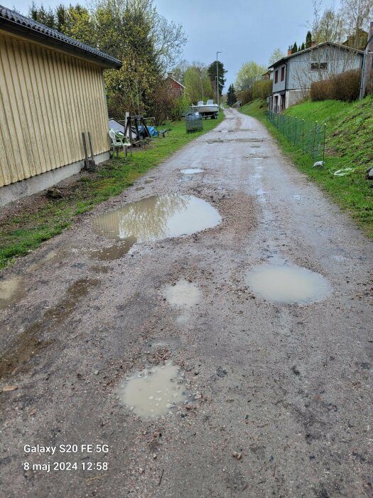 Grusväg med flera vattenpölar och omgivande bebyggelse samt vegetation i ett bostadsområde.