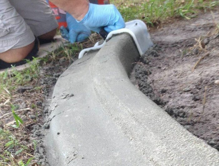 Person med blåa handskar formar nylagd betong på marknivå med hjälp av en plastform nära en gräsmatta.