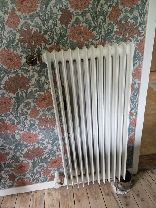 Vit vattenburen radiator monterad på en blommig tapet med en synlig rostfläck vid underkanten, stående på ett trägolv.