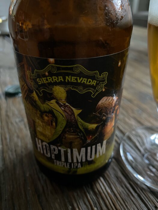 Flaska med etiketten "Sierra Nevada Hoptimum Triple IPA" på ett träbord, bredvid ett glas med öl.