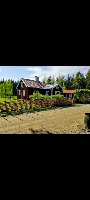 Röd stuga med vita knutar, zinkmagnesiumtak och en snygg skorsten. Omgiven av grönska med ett flätat trästaket och en grusväg framför huset.