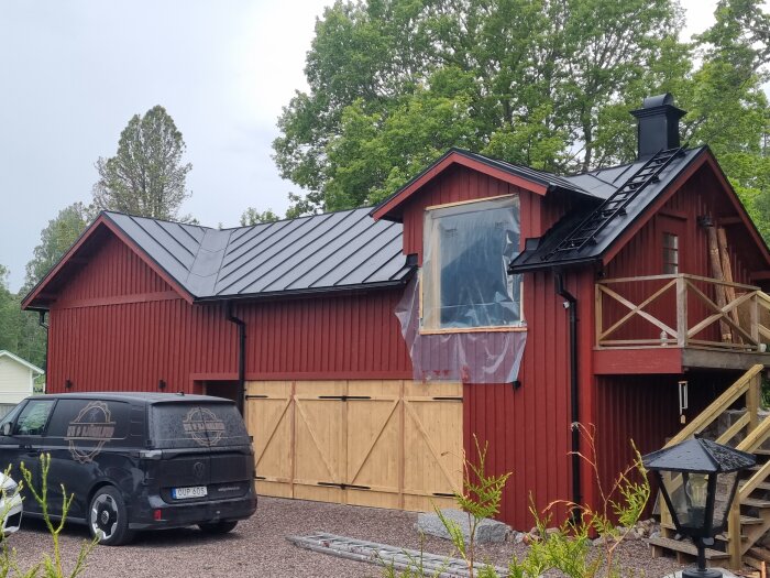 Rött hus med svarta zinkmagnesiumtak, pågående renovering. Ny skorsten och stora dörrar i trä. Svart skåpbil parkerad framför huset.