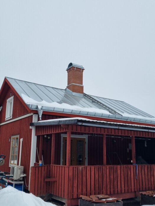 Rött hus med zinkmagnesiumtak, snötäckt skorsten och en veranda. Bilden visar exempel på tak- och skorstenarbete.