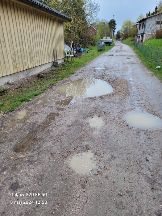 Grusväg med flera vattenpölar, vid sidan av vägen syns en gul byggnad, en plaststol och en båt täckt med presenning. Hus och träd i bakgrunden.