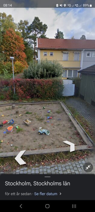 Sandlåda med leksaker framför gula hus med röd tak och omgärd av buskar och träd i ett bostadsområde.