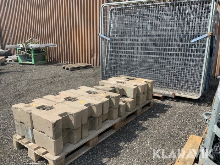 Staplade betongblock med slitsar på sidorna på en pall bredvid ett metallstaket på marken utanför en byggnad.