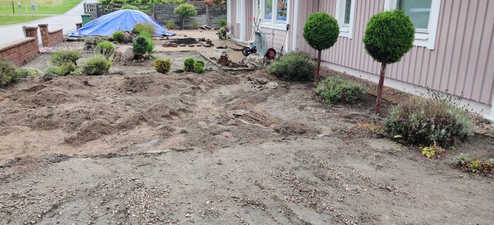 Trädgård med bortgrävt grus, kvarvarande sand i högar, några planterade buskar och ett blått tält i bakgrunden. Detaljerat arbete syns närmast huset.