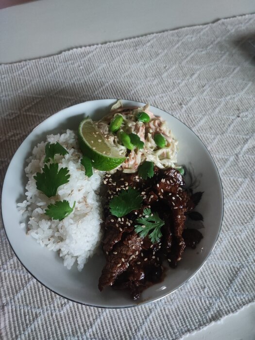 En tallrik med yakiniku, ris och en sallad garinerad med koriander och sesamfrön samt en limeklyfta.