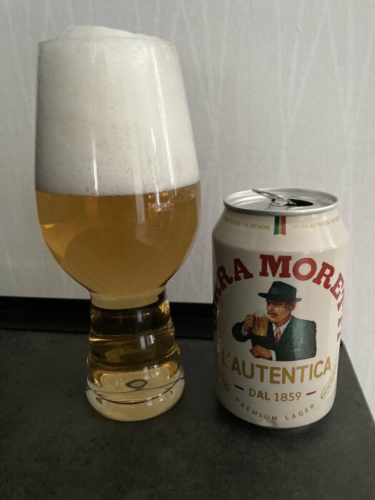 Ett glas öl med mycket skum bredvid en burk Birra Moretti L'Autentica premium lager.