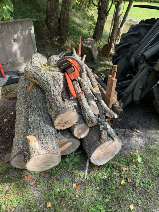 En hög med kapade trädstammar och kvistar ligger bredvid en motorsåg och en traktor i en skogsmiljö.