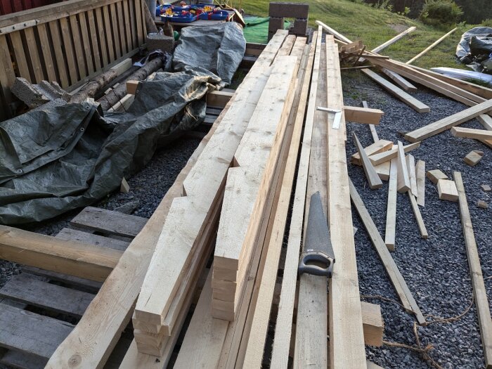 Högar av träbalkar och plankor ligger utspridda på en byggarbetsplats med en såg bland materialen, omgivna av grus och en presenning.