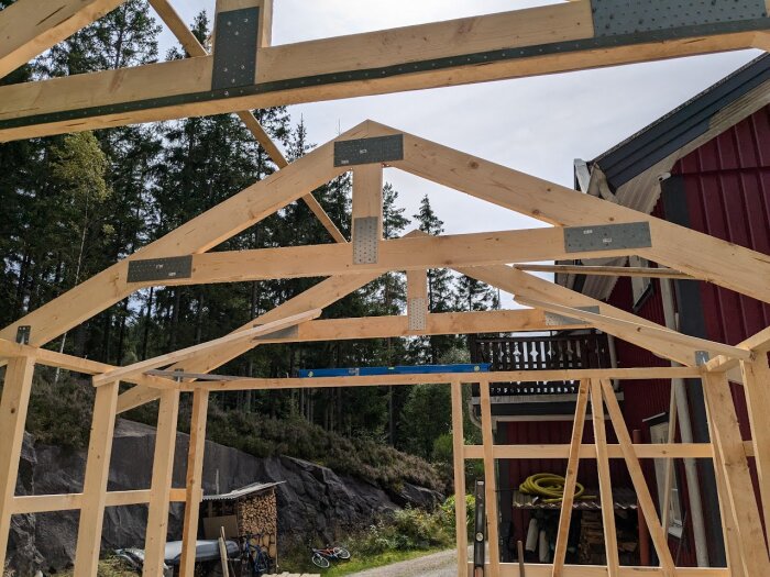 En stomme till ett tak av trä med takstolar spikade ihop med hålplattor och ankarspik under konstruktion, belägen nära en röd byggnad i skogslandskap.