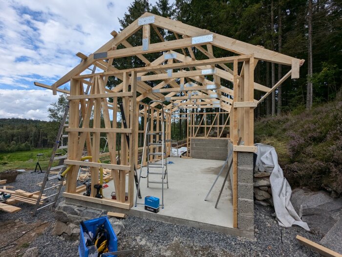 Trästomme med takstolar påloppsatta på en betongplatta under konstruktion, verktyg och byggmateriel synliga runtom, skog i bakgrunden.