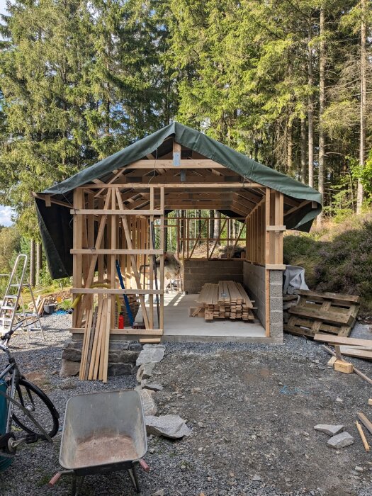 Pågående husbygge med trästomme och takstolar, täckt med en grön presenning. Stora träd i bakgrunden, byggmaterial och en skottkärra i förgrunden.