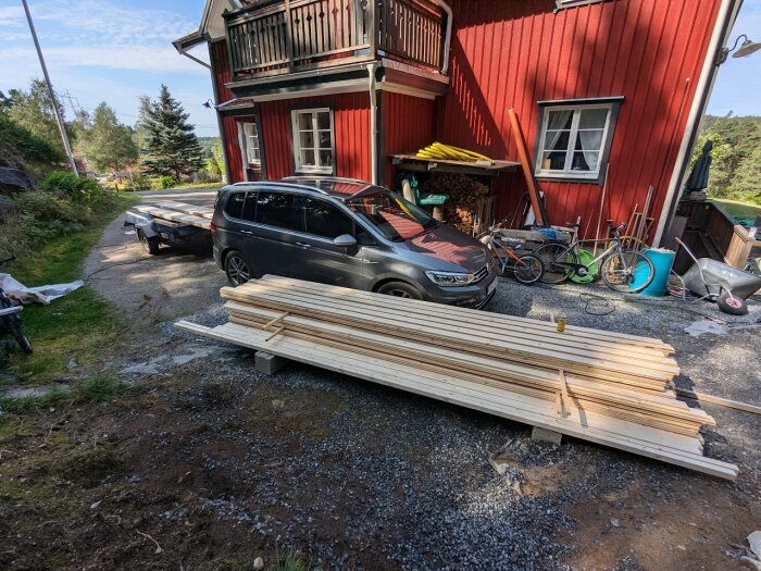 Staplade träbrädor på marken framför ett rött hus, med en svart bil parkerad nära och olika arbetsverktyg och cyklar i närheten.