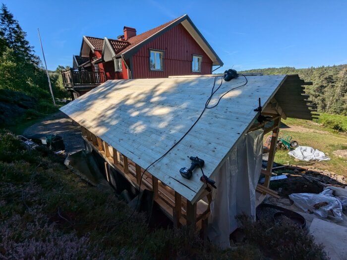 Bilden visar ett byggprojekt där råsponten för ett tak är nästan färdiglagt på en byggnad. Diverse verktyg ligger på taket, inklusive en borrmaskin.