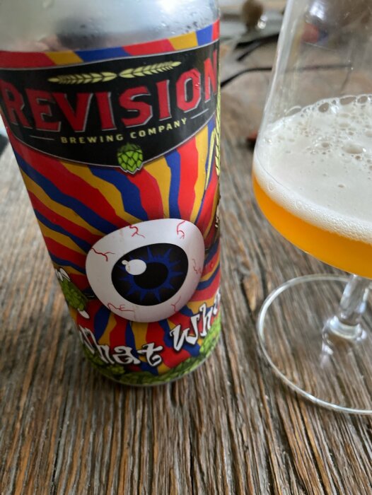 Ölburk från Revision Brewing Company och ett glas med öl på ett träbord. Etiketten har en färgglad design med ett stort blått öga.