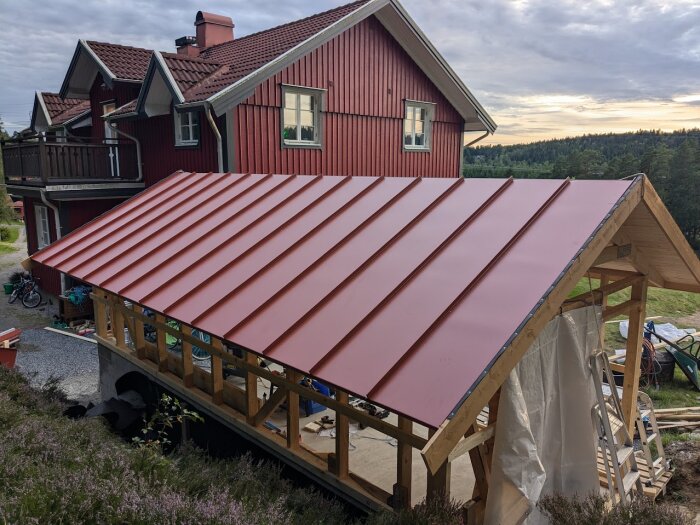 Rött hus med nytt rött klicktak i närbild, med landskap och skog i bakgrunden. Byggnadens träkonstruktion synlig under taket.