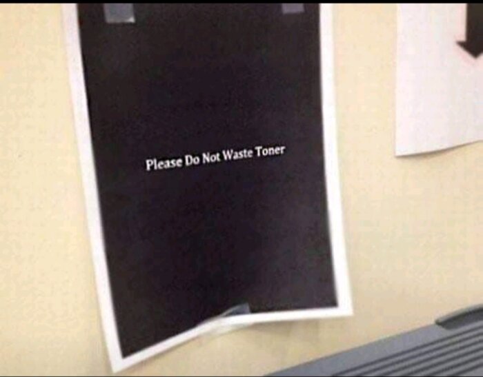 Bild på ett papper med texten "Please Do Not Waste Toner" tryckt mot en svart bakgrund, upphängt på en vägg nära en skrivare.