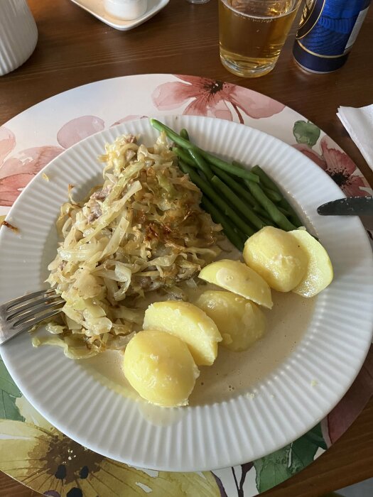 Tallrik med kålpudding, potatis, haricots verts och gräddsås på ett underlägg med blommönster, med en dryck i glas och ölburk i bakgrunden.