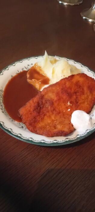 En tallrik med schnitzel, potatismos, madeirasås och en klick crème fraiche på ett bord.