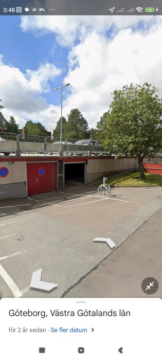 Parkeringsplats ovanför garageportar under bro med sittföremål och skyltar på väggar, träd på gräsmatta bredvid, klarblå himmel med moln i bakgrunden.