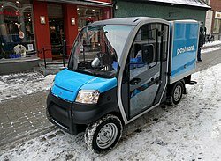 Blå och vit Postnord transportbil parkerad på en snötäckt gata framför några butiker.