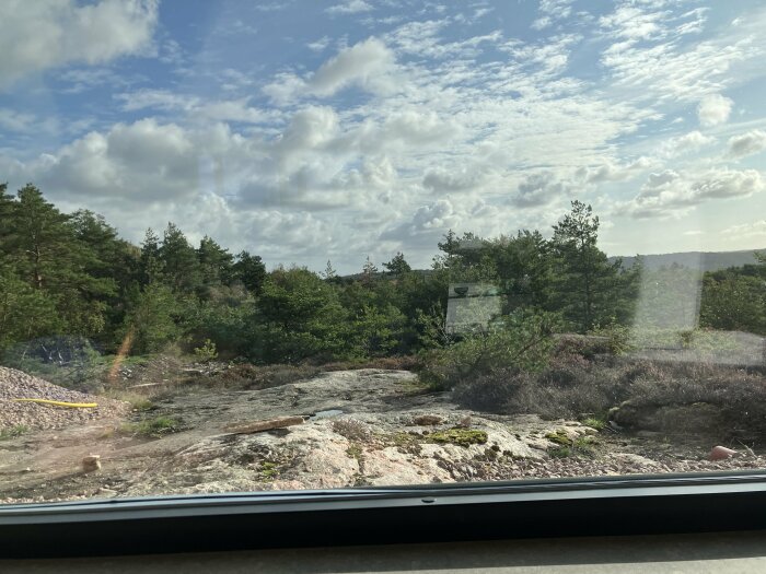 Utsikt genom ett fönster där man ser en stenig skogsslänt med träd under en molnig himmel. Fönstrets nederkant synlig med reflektioner.