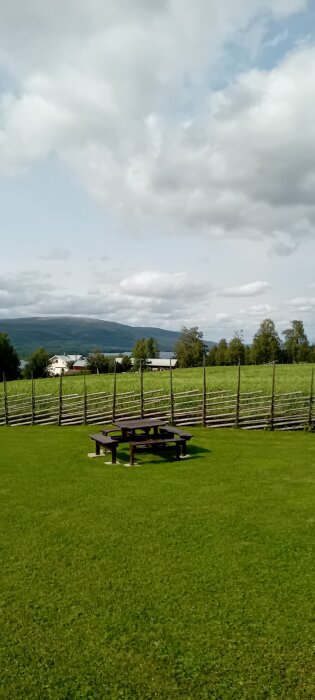 Picknickbord och bänkar på en grön gräsmatta med en trästaket i bakgrunden, omgivna av öppna fält, träd och berg i fjärran.