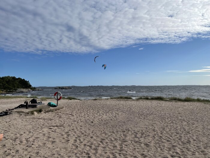 Strandbild med livboj och kitesurfare i blåsig miljö.