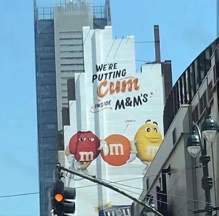 Stor reklamskylt på en byggnad i stadsmiljö som reklamerar för M&M's godis, men med en felstavning i texten där "sum" har blivit "cum".