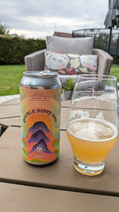 En burk med ölet Double Dippy Doo Hazy DIPA från Dry & Bitter Brewing Company och ett glas med samma öl. Offentlig plats med trädgård i bakgrunden.