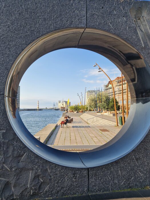 Utsikt genom ett cirkulärt monument över en hamn med en fyr i bakgrunden, människor som sitter på bänkar längs kajen och byggnader till höger.