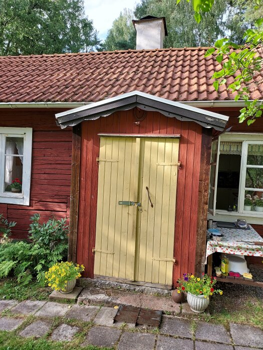 Rött trähus med tegeltak och dubbelgula dörrar med svart lås. Två blomkrukor med gula och flerfärgade blommor framför dörren.