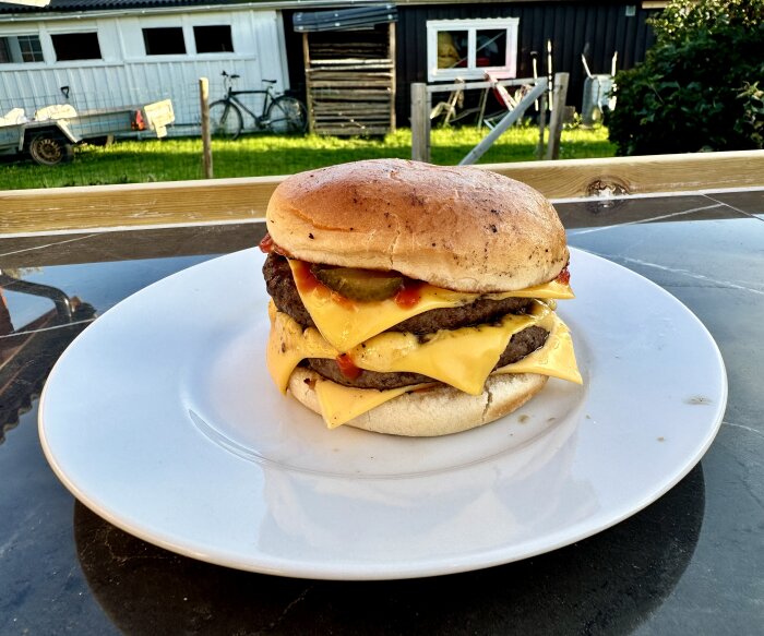 En dubbelcheeseburgare med pickles och ketchup på en vit tallrik utomhus, med en trädgård och byggnader i bakgrunden.