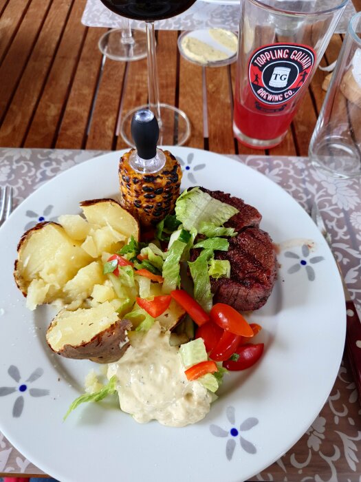 En tallrik med bakad potatis, grillad oxfilé, majs, grönsaker och bearnaisesås, ett vinglas och ett ölglas i bakgrunden på ett träbord.