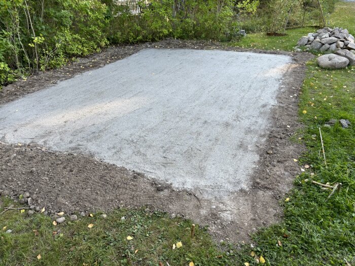 Nyutjämnad yta med grus och paddning förberedd för läggning av marksten, omgiven av gräs och träd. En hög med större stenar syns i bakgrunden.