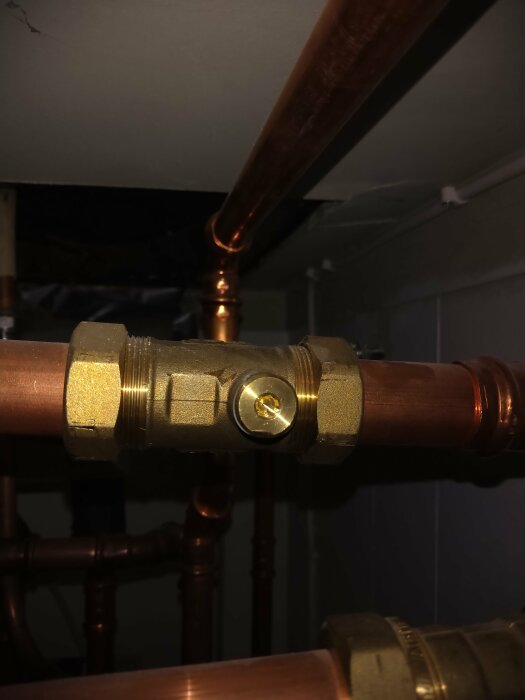 Närbild på en metallventil eller koppling monterad på ett kopparrör, troligen en del av ett värmesystem, ovanför innedelen av en värmepump.