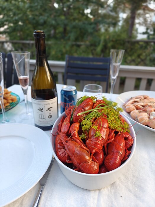 Skål med kräftor på ett dukat bord tillsammans med en vinflaska och en sodaburk, omgiven av tallrikar och glas i utomhusmiljö.