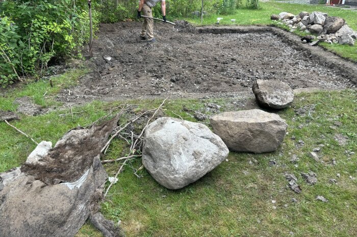 Person gräver i en jordig tomt med rötter och stora stenar i förgrunden. Ett pågående byggprojekt i en trädgård med grönska runt omkring.