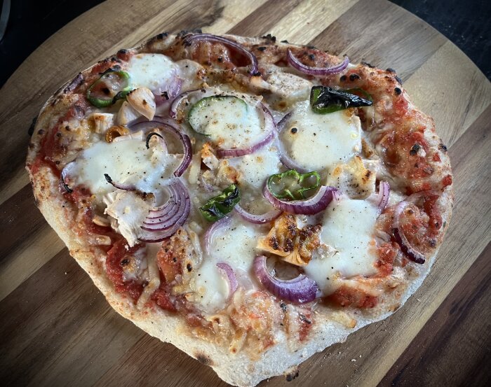 Hemgjord pizza med tomatsås, mozzarella, grillad kyckling, rödlök och padron på en träskärbräda.