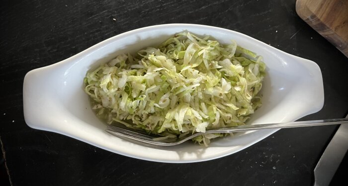 Coleslaw med salladslök och rödlök i en vit serveringsskål med en gaffel, placerad på ett mörkt bord.