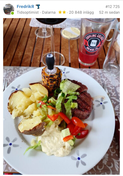 Tallrik med kött, potatis, sallad och sås, tillsammans med majs på en pinne och ett glas med röd saft på ett träbord.