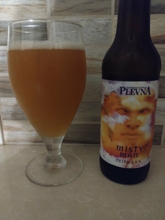 En flaska Plevna Misty Man NEIPA 5,5% öl bredvid ett glas med samma öl, fotograferade mot en kaklad vägg.