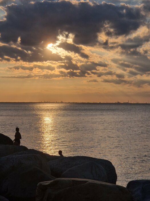 Solnedgång över havet med två silhuetter av människor som står på stenar vid vattenkanten, mörka moln på himlen och reflektioner i vattnet.